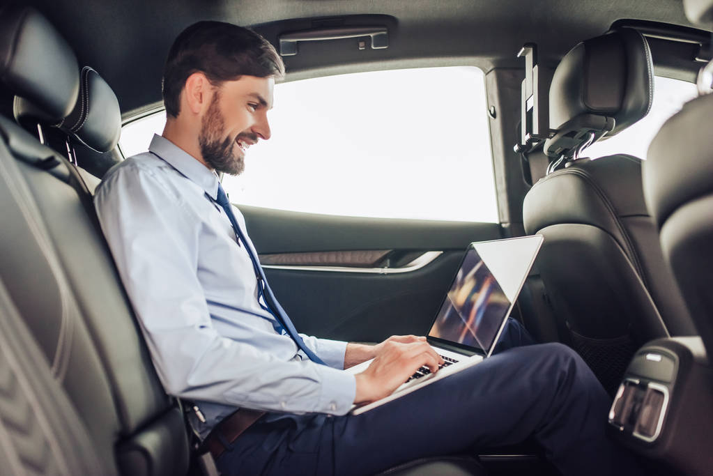 vista lateral del joven hombre de negocios barbudo que trabaja en el ordenador portátil en el coche
 - Foto, Imagen