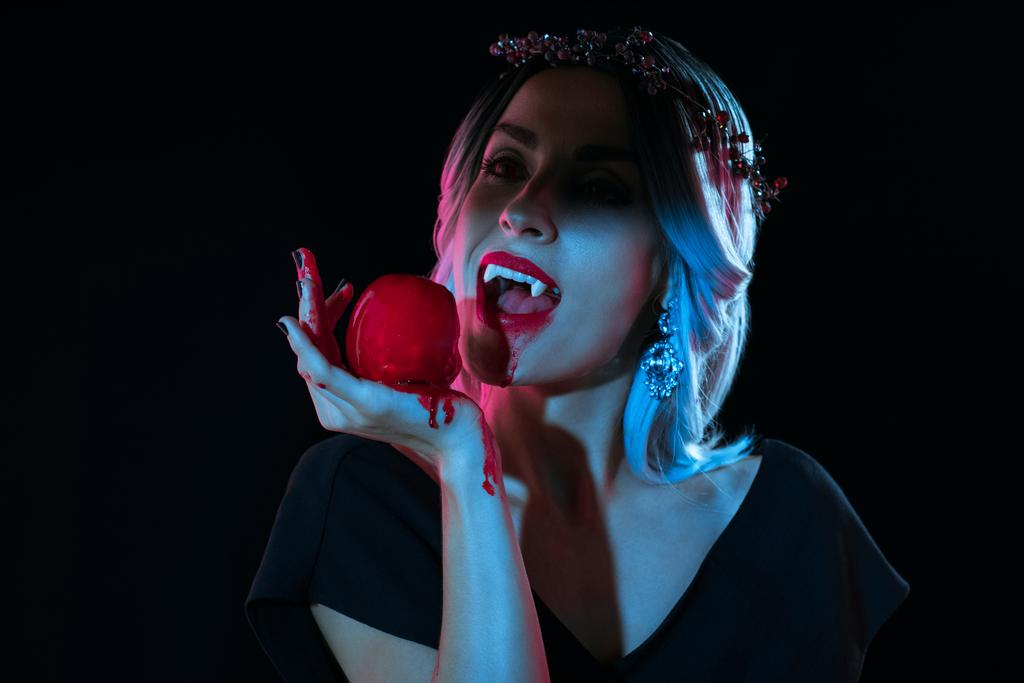 attractive vampire woman bitting red bloody apple isolated on black - Photo, Image