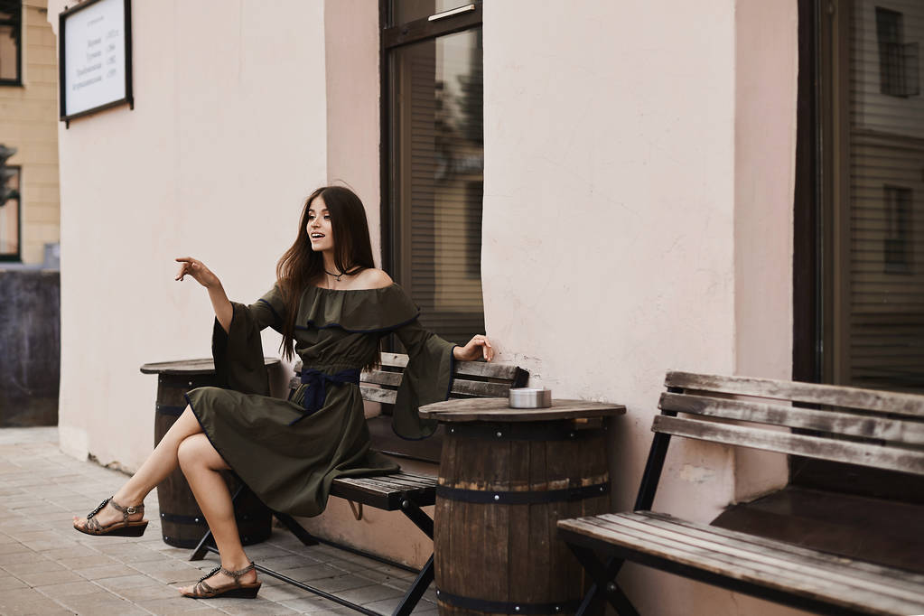 Krásná a elegantní brunetka model dívka ve stylových šatech s nahá ramena sedí na lavičce a pózuje venku na malé náměstí - Fotografie, Obrázek