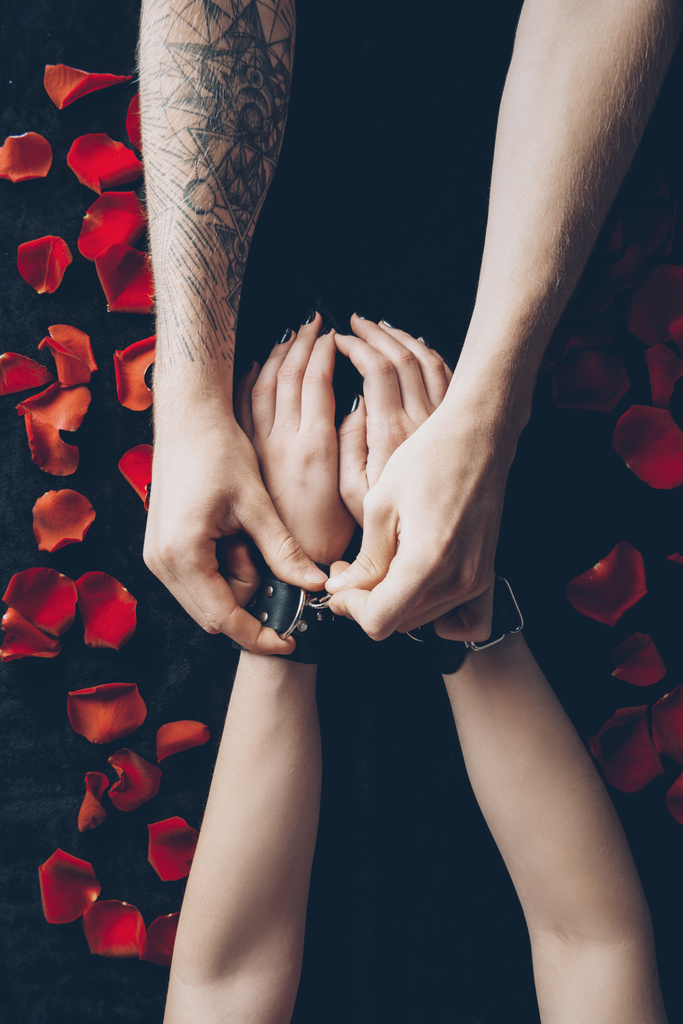 обрізаний знімок пари зі шкіряними наручниками над чорною тканиною з пелюстками червоної троянди
 - Фото, зображення