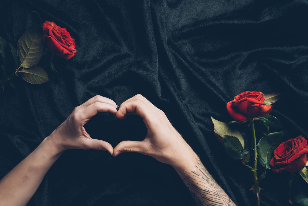 Schnappschuss eines Paares, das Hand-Herz-Symbol über schwarzem Stoff mit roten Rosen zeigt - Foto, Bild