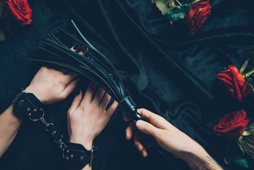 обрізаний знімок чоловіка, що тримає чорний батіг і жінку в шкіряних наручниках
 - Фото, зображення