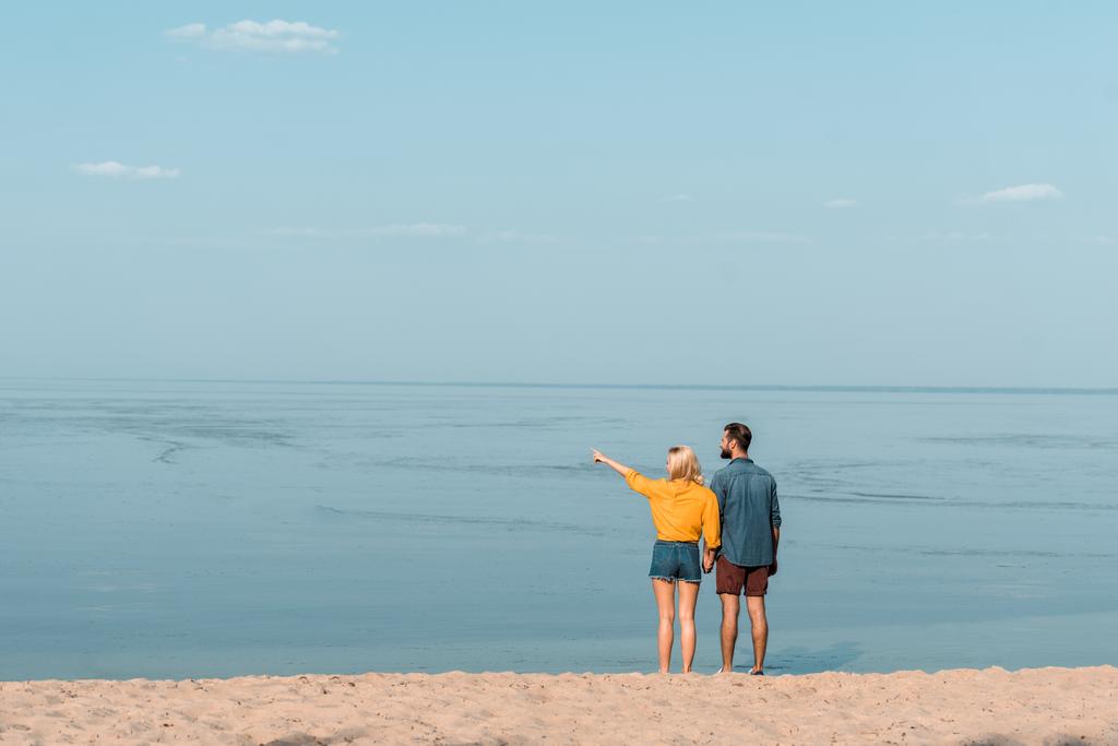 Rückansicht des Paares Händchen haltend am Strand, Freundin zeigt auf etwas - Foto, Bild
