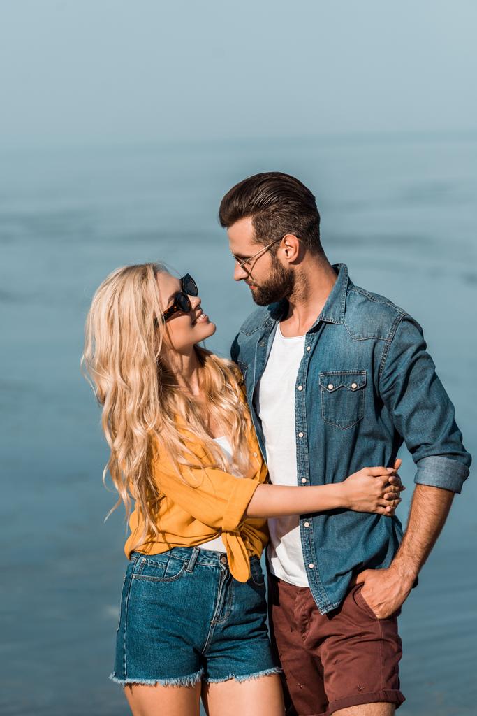 pareja en gafas de sol abrazándose y mirándose cerca del mar
 - Foto, imagen