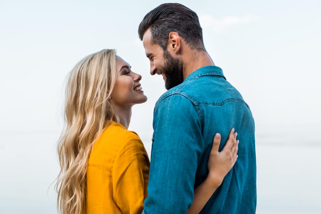 vista lateral de la pareja sonriente abrazándose y mirándose el uno al otro contra el cielo azul
 - Foto, imagen