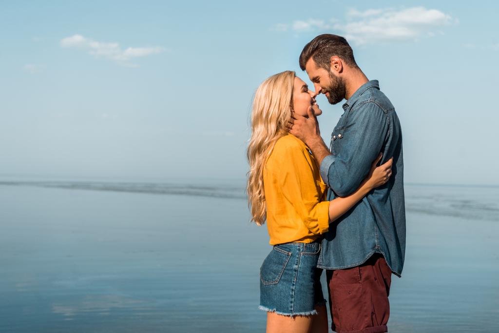 side view of smiling girlfriend and boyfriend touching with noses on beach - Photo, Image