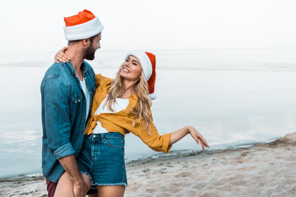 пара в шляпах Санта-Клауса обнимаются и веселятся на пляже
 - Фото, изображение