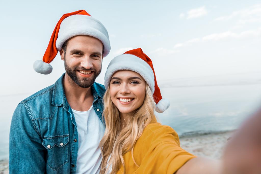camera point of view of smiling couple in santa hats looking at camera on beach - Photo, Image