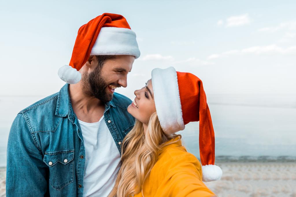 sorridente coppia in cappelli di Babbo Natale abbracciarsi sulla spiaggia e guardarsi l'un l'altro
 - Foto, immagini