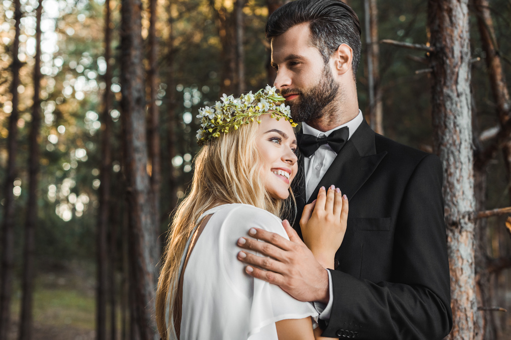schöne lächelnde Braut im weißen Kleid und schöner Bräutigam im Anzug, der sich im Wald umarmt  - Foto, Bild