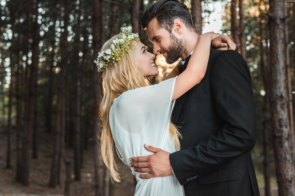 beautiful happy bride in white dress and handsome groom in suit hugging and going to kiss in forest  - Photo, Image