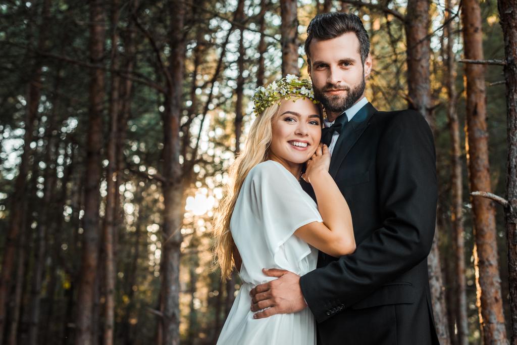 Tiefansicht eines lächelnden Hochzeitspaares, das sich umarmt und bei Sonnenuntergang im Wald in die Kamera schaut - Foto, Bild