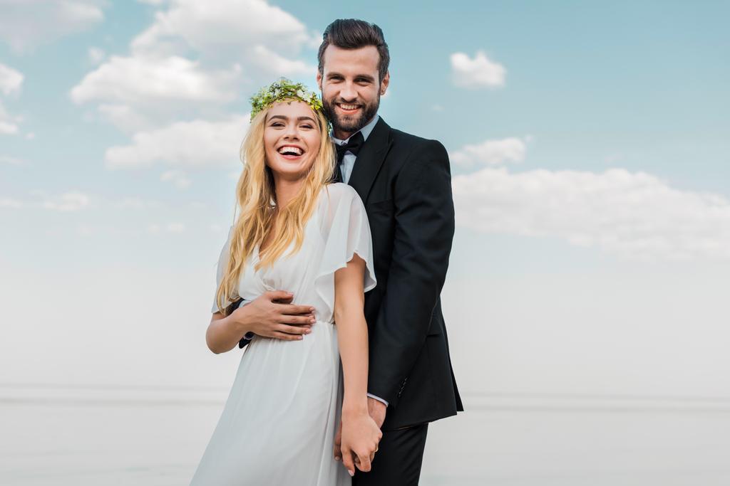 Смеющаяся свадебная пара в костюме и белом платье, держащиеся за руки и смотрящие в камеру на пляже
 - Фото, изображение