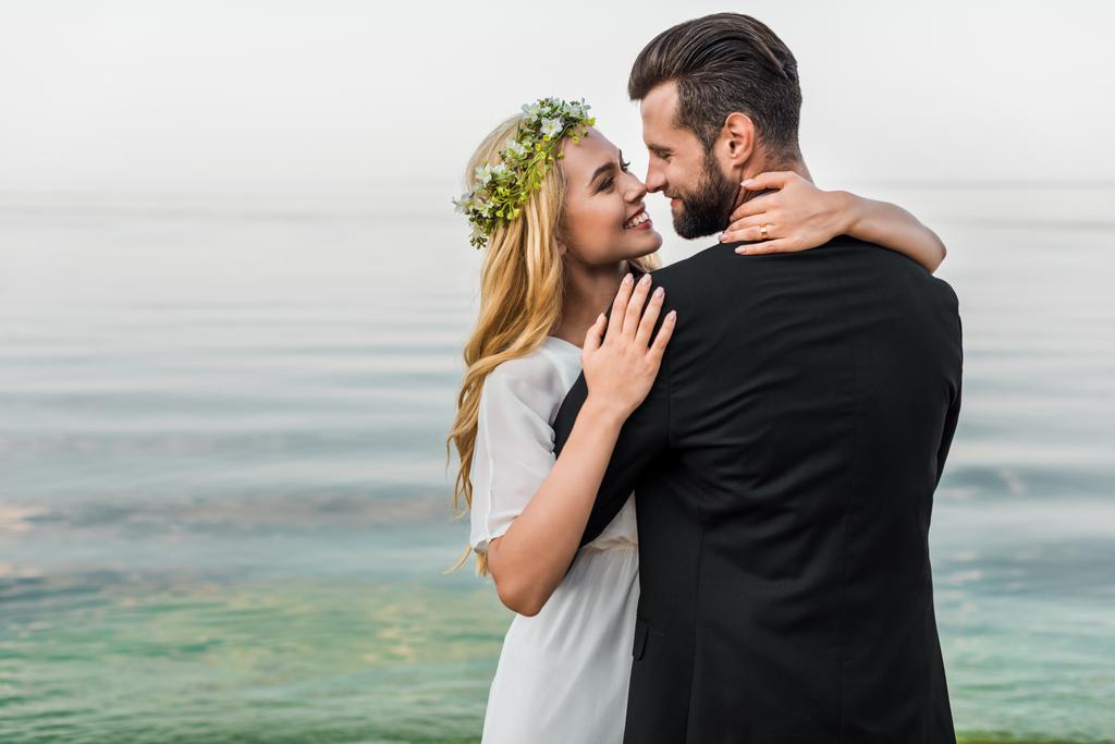 atractiva novia en guirnalda y novio en traje abrazándose en la playa y tocando con la nariz
 - Foto, imagen