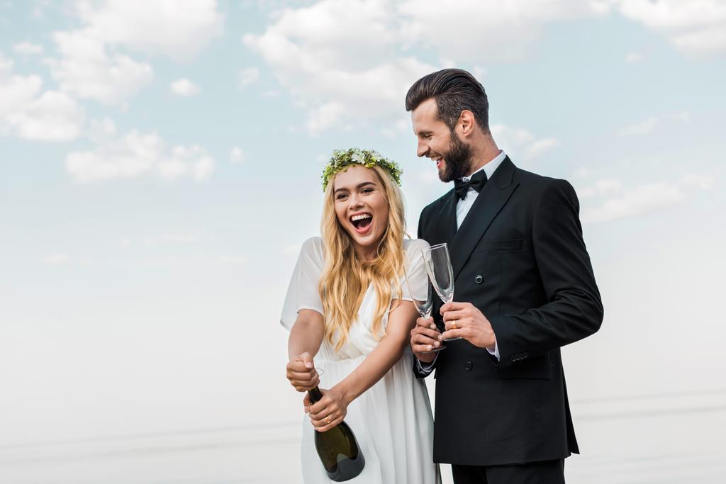 gelukkige bruid in witte jurk openen champagnefles op strand - Foto, afbeelding