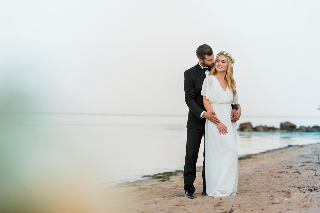 γαμπρός στο κοστούμι αγκάλιασμα ελκυστική νύφη σε λευκό φόρεμα στην παραλία - Φωτογραφία, εικόνα