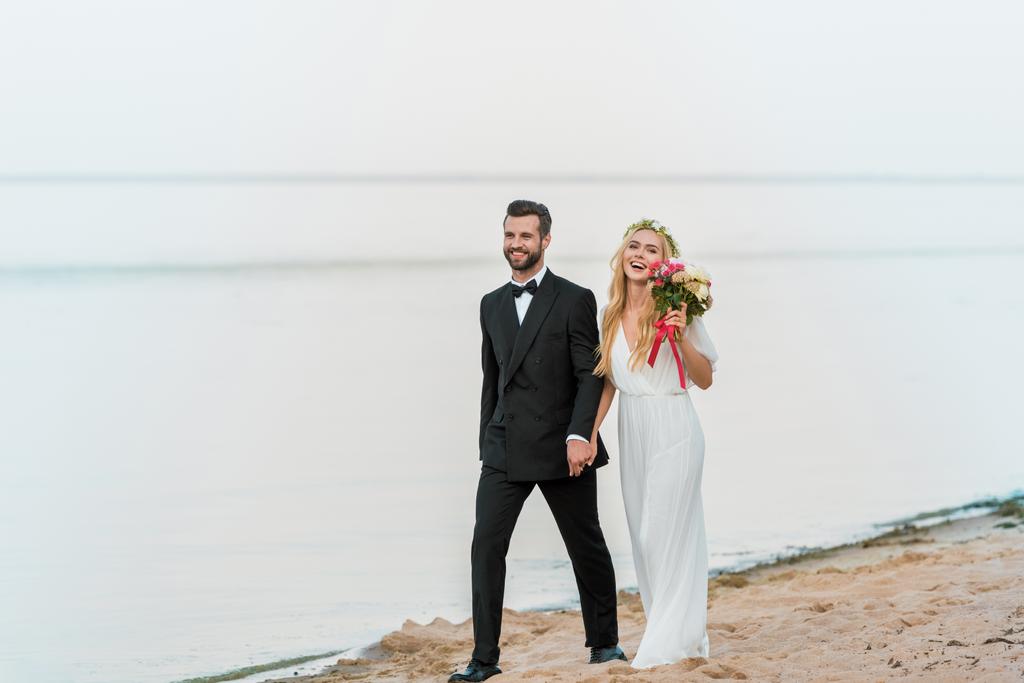 χαμογελώντας γαμήλιο ζεύγος κρατώντας τα χέρια και τα πόδια στην παραλία - Φωτογραφία, εικόνα