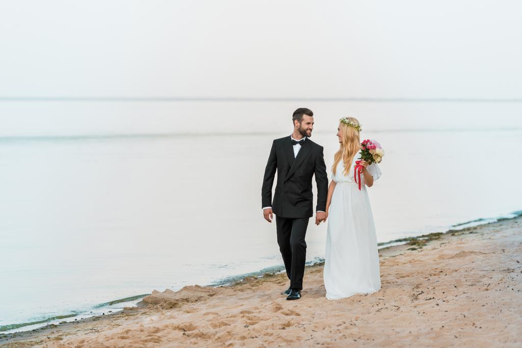 Svatební pár drží za ruce, chůzi a při pohledu na sebe na pláži - Fotografie, Obrázek