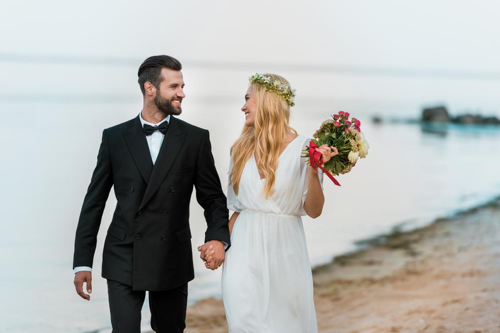affectionate wedding couple holding hands, walking and looking at each other on beach - Photo, Image