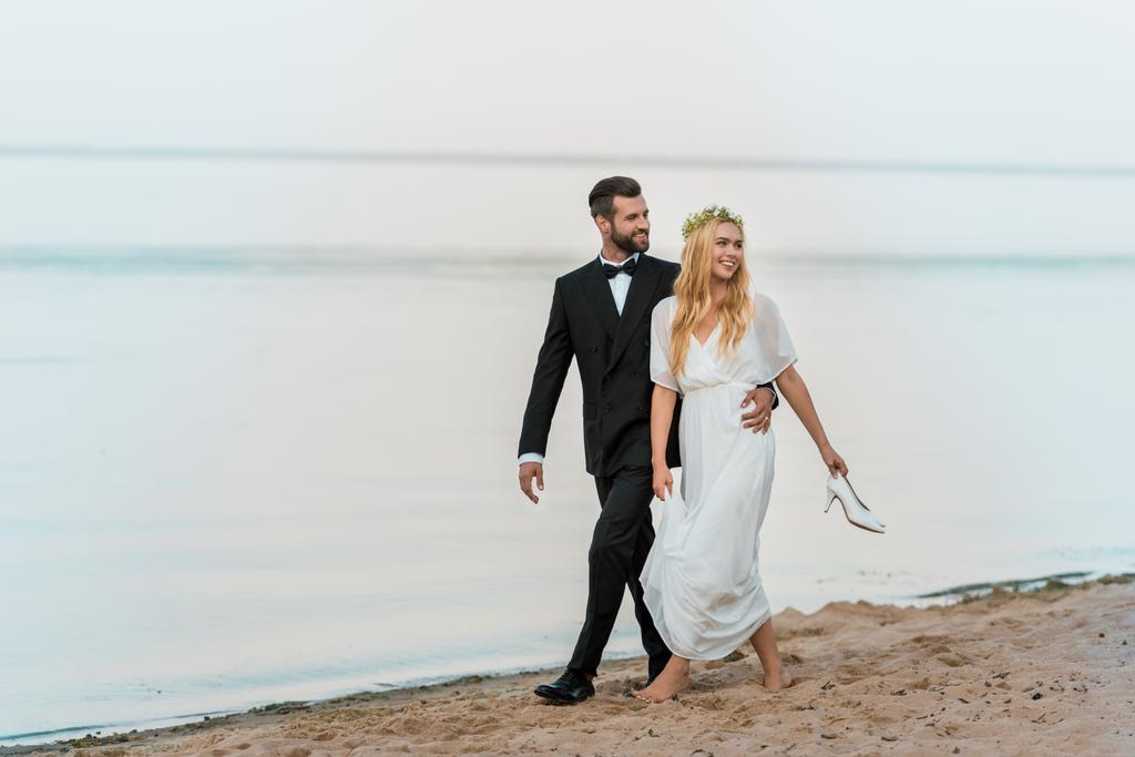 свадебная пара обнимается, гуляет по пляжу и смотрит в сторону, невеста держит высокие каблуки в руке
 - Фото, изображение