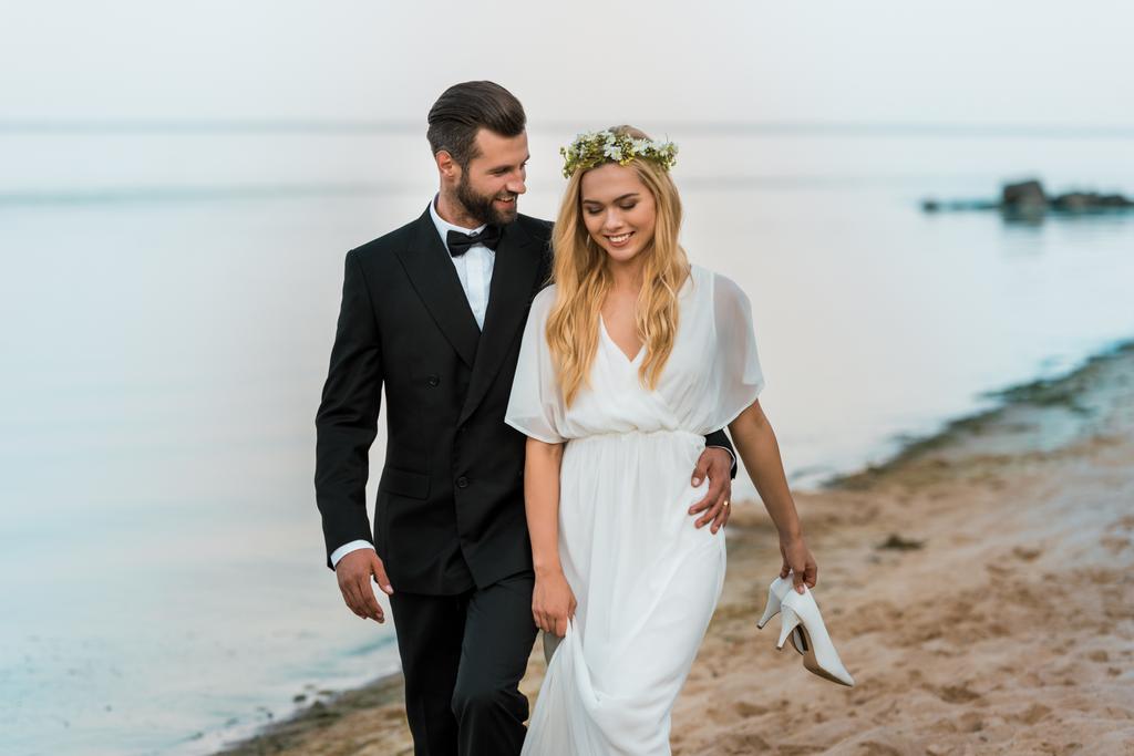 milující svatební pár objímat a procházky na pláži, nevěsta v ruce drží vysoké podpatky - Fotografie, Obrázek