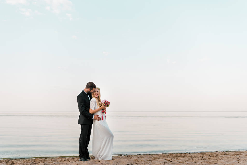 ženicha objímání nevěsta a ona drží svatební kytice na písčité mořské pláži - Fotografie, Obrázek