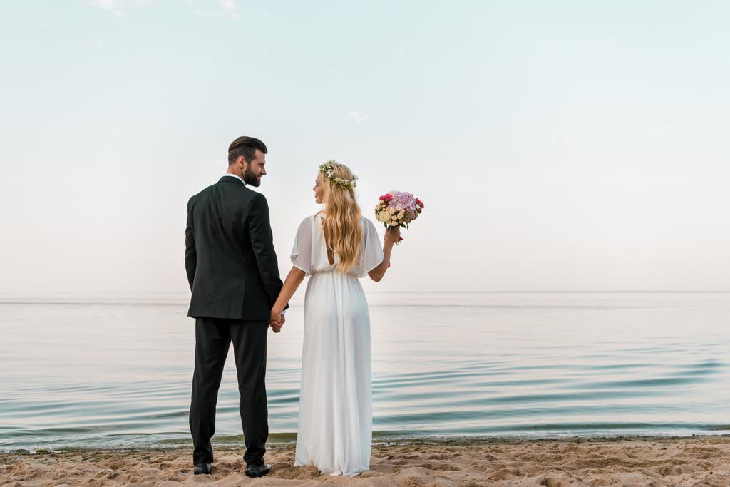 vista trasera de la pareja de boda de pie en la playa con ramo de bodas y mirándose entre sí
 - Foto, imagen