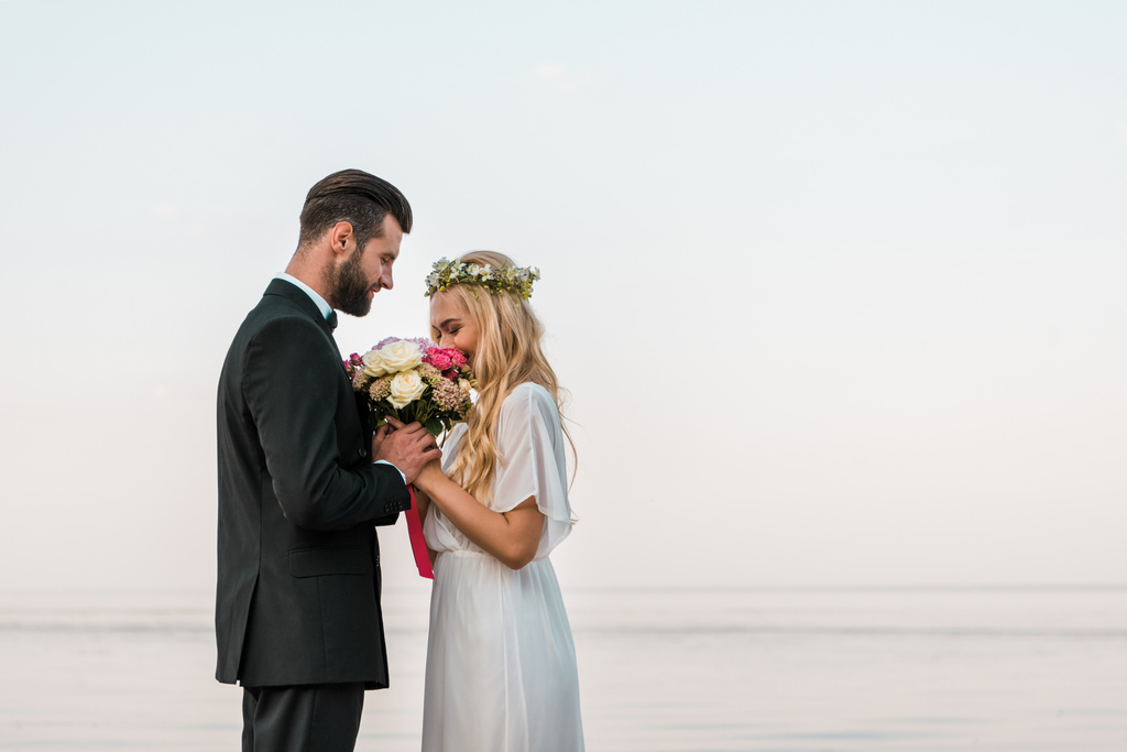 Seitenansicht des Hochzeitspaares mit Blumenstrauß am Strand, Braut schnüffelt an Rosen - Foto, Bild