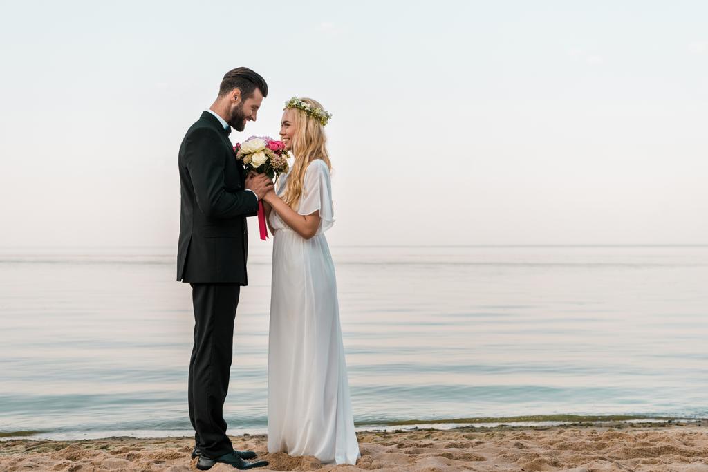 vista lateral de pareja de boda cariñosa de pie con ramo en la playa, novia oliendo rosas
 - Foto, imagen