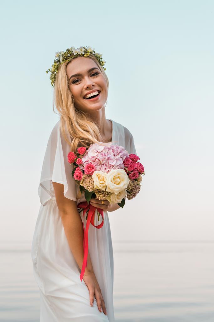γελώντας ελκυστική νύφη σε λευκό φόρεμα και στεφάνι κρατώντας γαμήλια ανθοδέσμη στην παραλία και βλέπουν τα φωτογραφικών μηχανών - Φωτογραφία, εικόνα