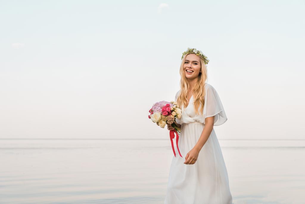 novia atractiva sonriente en vestido blanco y corona celebración de ramo de boda y caminar cerca del mar
 - Foto, imagen