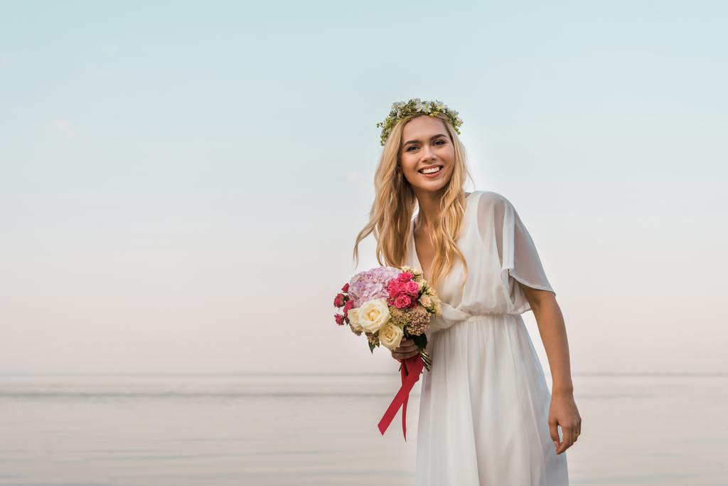 beyaz elbise ve düğün buket gül tutan ve plaj kameraya bakarak çelenk içinde çekici gelin gülümseyen - Fotoğraf, Görsel