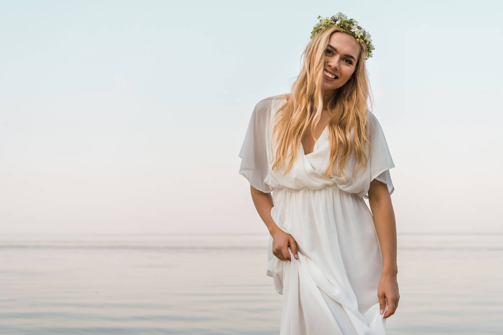 uśmiechający się atrakcyjna Panna Młoda w białej sukni i Wieniec kwiatów, patrząc na kamery w pobliżu ocean - Zdjęcie, obraz
