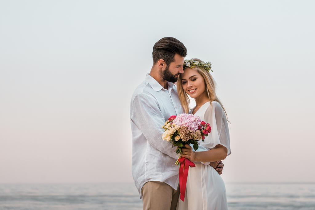 bride with wedding bouquet and groom hugging on beach - Photo, Image