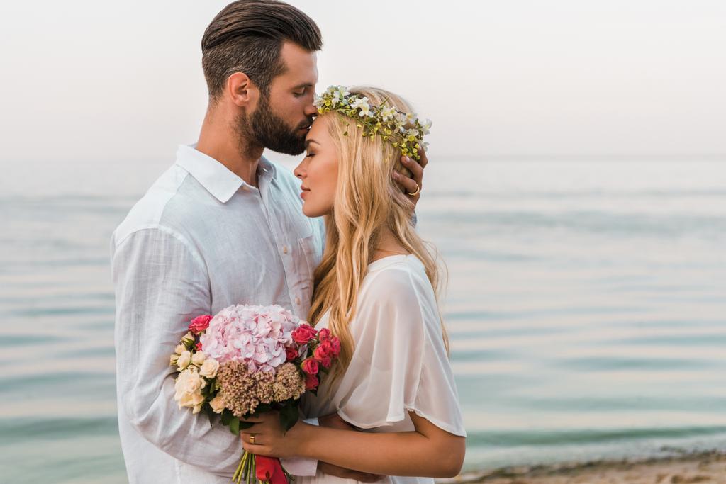 vista lateral do belo noivo beijar noiva testa na praia
 - Foto, Imagem