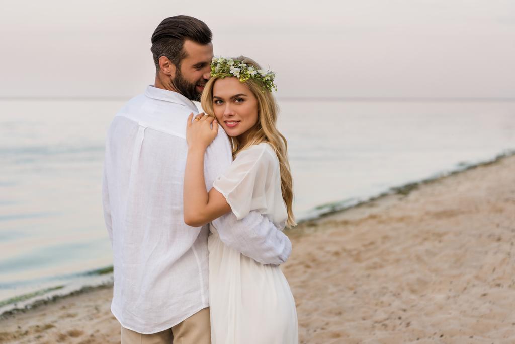 Bräutigam umarmt attraktive Braut im Kranz und sie blickt in die Kamera am Strand - Foto, Bild