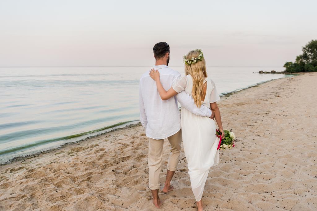 Achteraanzicht van de bruid met bruiloft boeket en bruidegom knuffelen en wandelen op strand - Foto, afbeelding