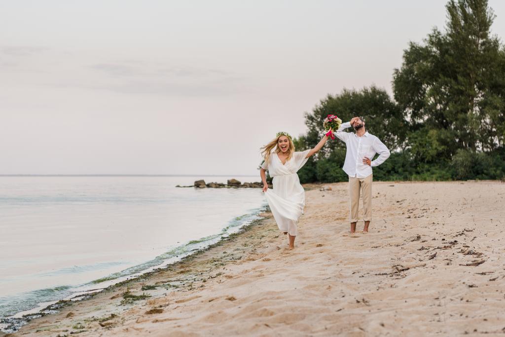 šťastná nevěsta běží s svatební kyticí, ženich šklebící se na pláži - Fotografie, Obrázek