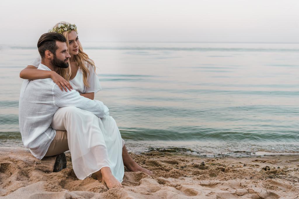 eleganter Bräutigam und Braut sitzen auf Baumstämmen am Strand und schauen weg - Foto, Bild
