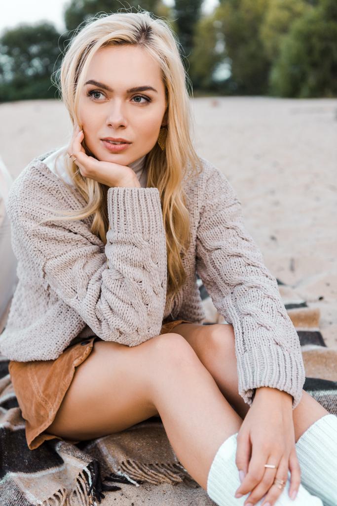 attractive pensive blonde girl in autumn outfit sitting on beach and looking away - Photo, Image