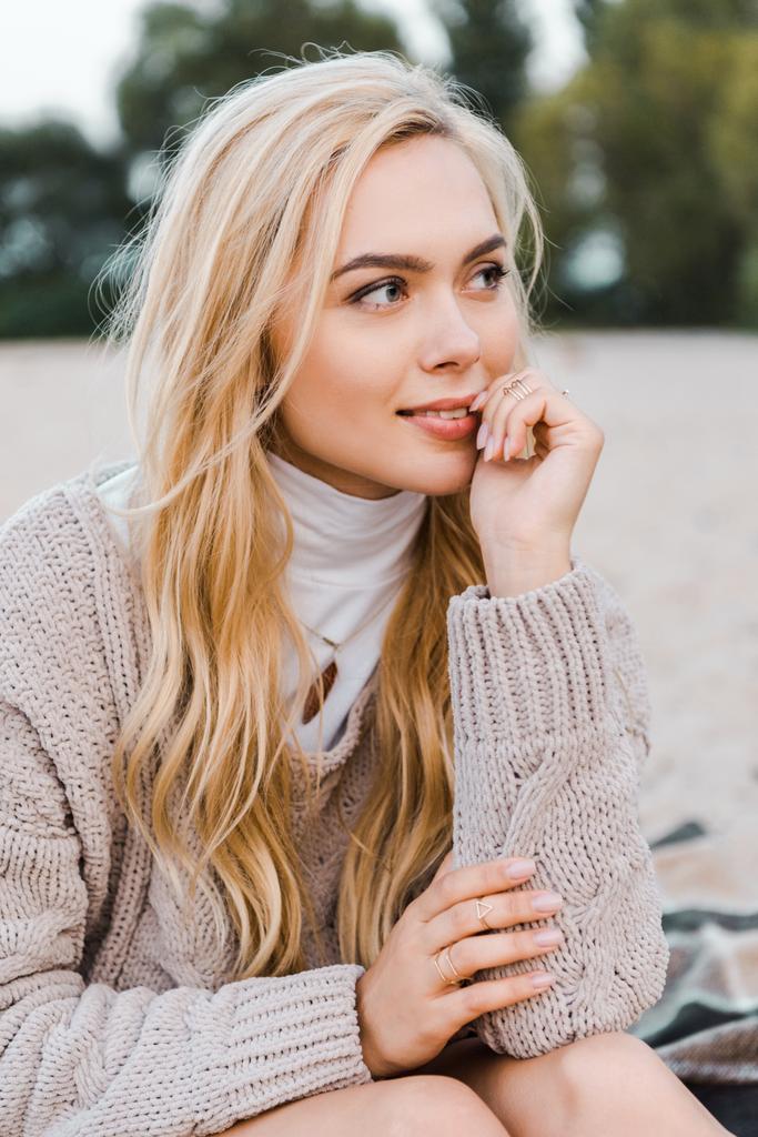 attractive smiling blonde girl in autumn outfit sitting on beach, touching lips and looking away - Photo, Image
