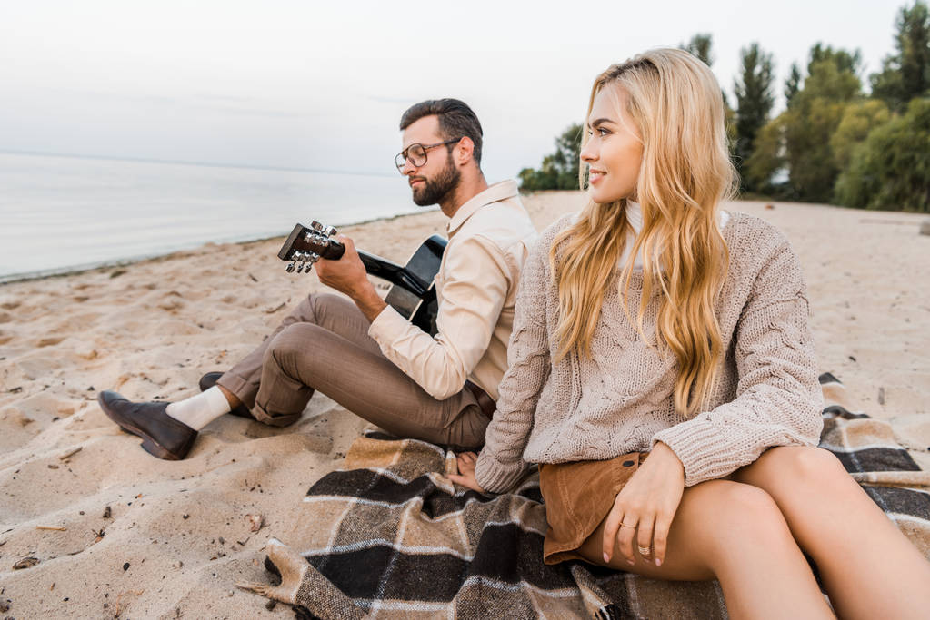 handsome boyfriend in autumn outfit playing acoustic guitar for girlfriend on beach - Photo, Image