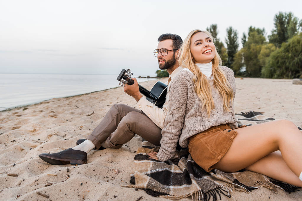 handsome boyfriend in autumn outfit playing acoustic guitar for smiling girlfriend on beach - Photo, Image