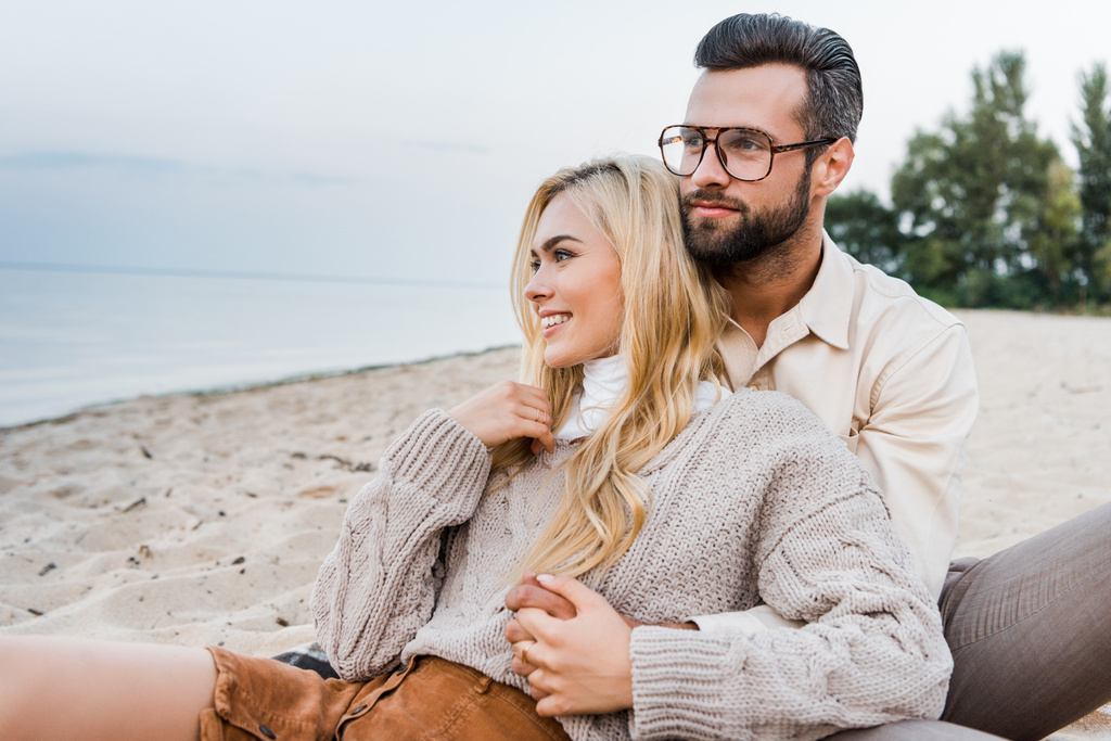 fröhliches Paar im Herbst-Outfit sitzt und umarmt sich am Strand - Foto, Bild