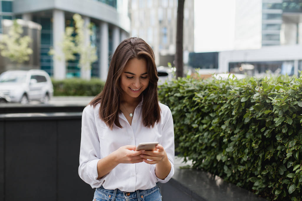 Ευτυχής επιχειρηματίας χρησιμοποιώντας κινητό τηλέφωνο κοντά στο γραφείο, όμορφη γυναίκα περιήγησης τηλεφώνου χαμογελώντας περπάτημα σε εξωτερικούς χώρους, θηλυκό διαχείριση γραπτών μηνυμάτων σε μια εφαρμογή messenger χρησιμοποιώντας σύγχρονο smartphone  - Φωτογραφία, εικόνα