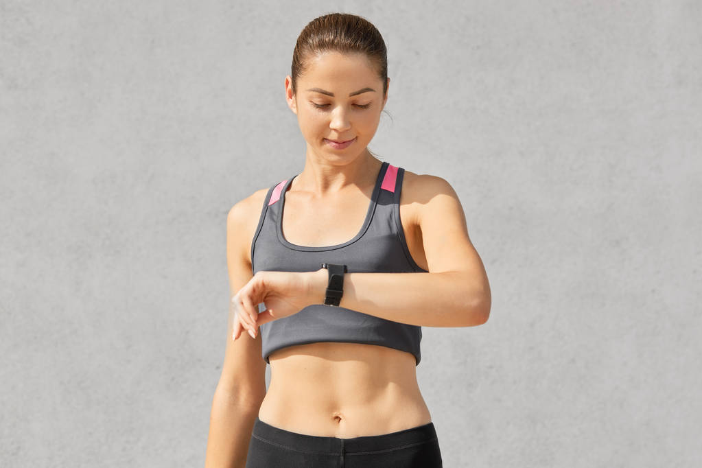 Shot of fitness lady or jogger looks attentively at smart watch, checks pulse and counts calories, has healthy lifestyle, goes in for sport regularly, dressed in casual tanktop, shows perfect figure - Photo, Image