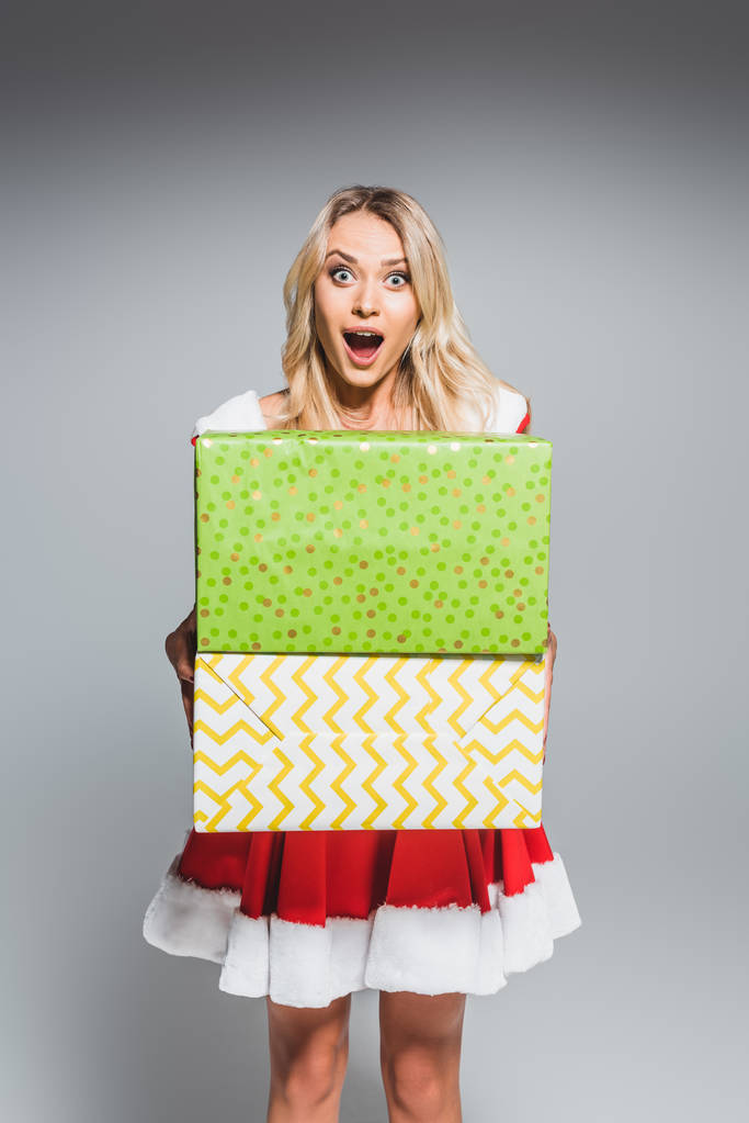 chocado bela jovem mulher no vestido de Natal segurando caixas de presente isolado no fundo cinza
 - Foto, Imagem