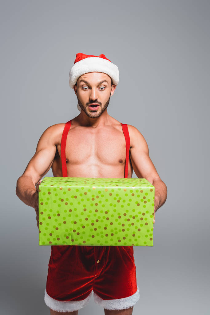 schockierter muskulöser Mann mit Weihnachtsmütze hält Geschenkbox isoliert auf grauem Hintergrund - Foto, Bild