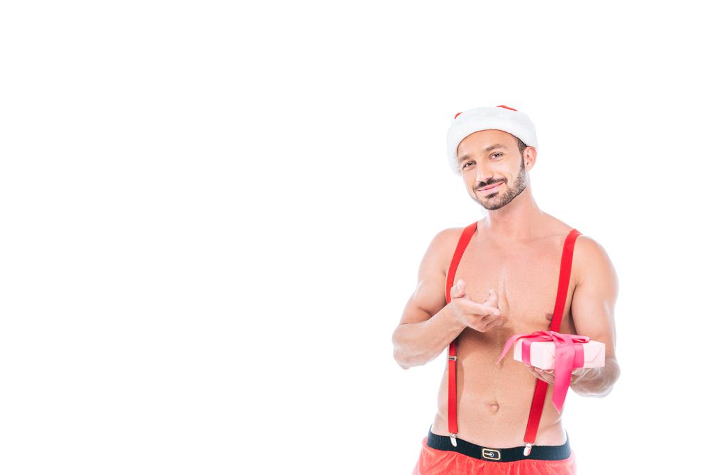 щасливий м'язистий чоловік у різдвяному капелюсі, що вказує на подарункову коробку ізольовано на білому тлі
 - Фото, зображення