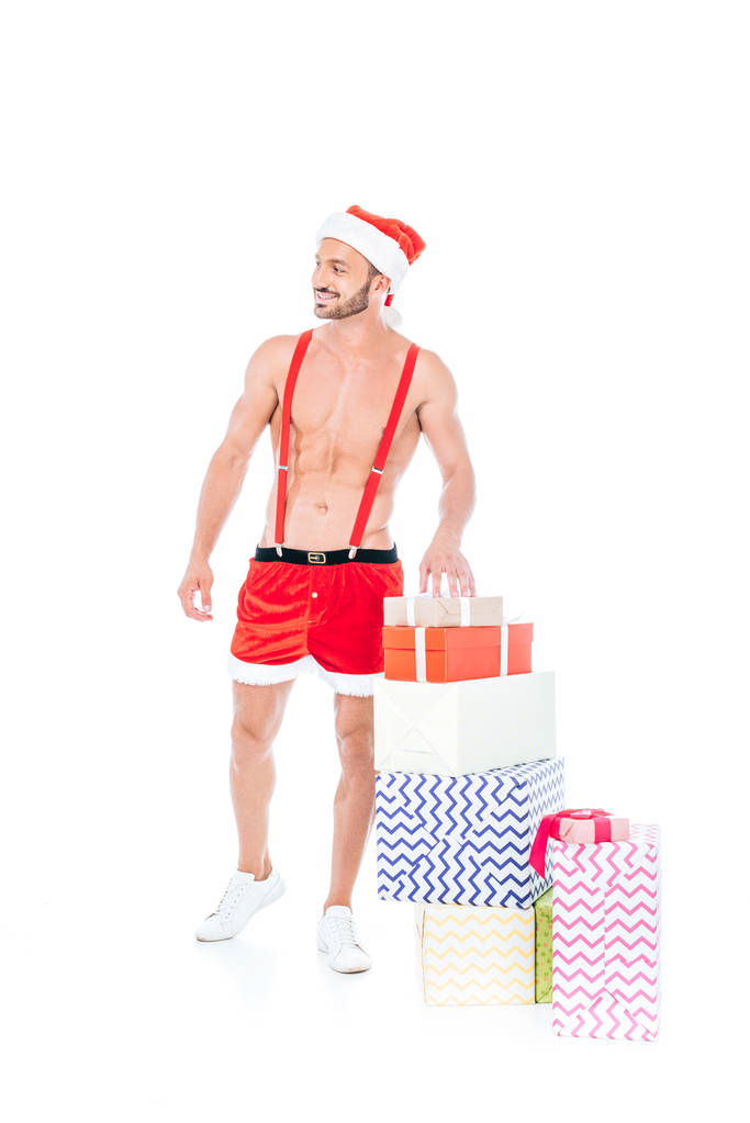 shirtless μυώδης άνδρας σε Χριστουγεννιάτικο καπέλο στέκεται με σωρό κουτιά δώρων που απομονώνονται σε λευκό φόντο - Φωτογραφία, εικόνα